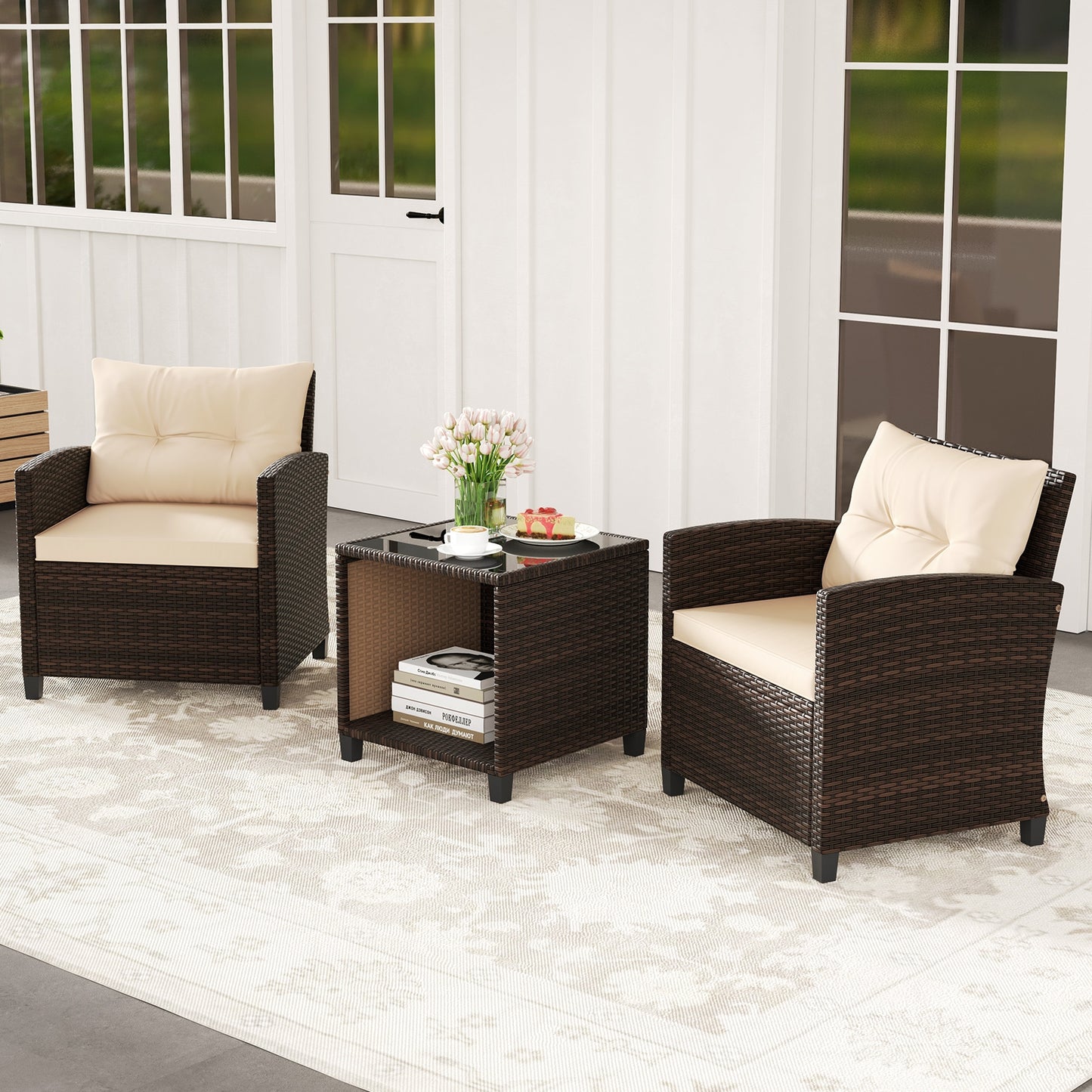A three piece patio sofa set with two wicker chairs and a coffee table with a storage compartment. The set is beige and brown in color with cushions on the chairs and a book and a vase of flowers on the table, placed on a white rug.