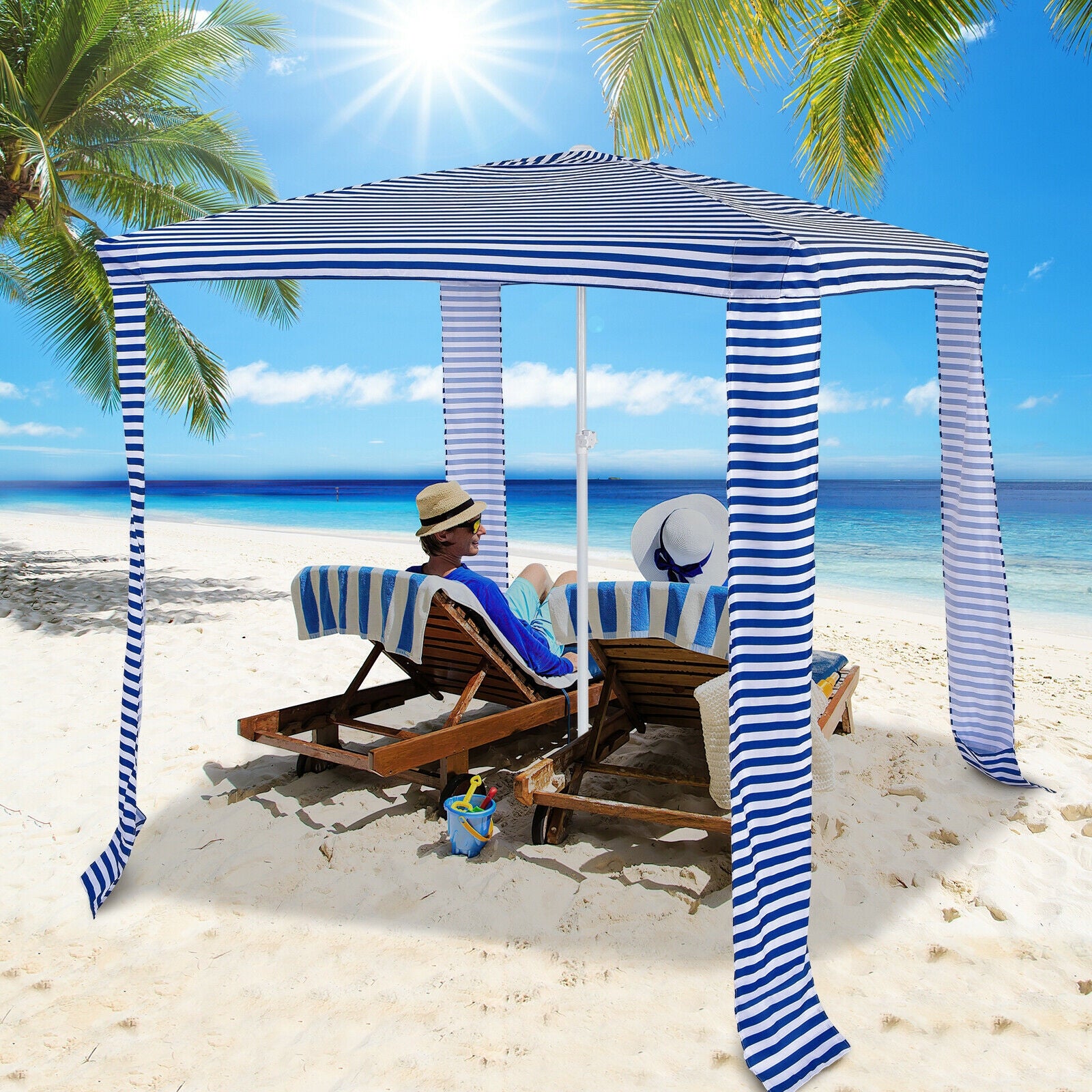 A navy and white striped beach canopy set up on a beach with a person lounging on a chair under it, surrounded by palm trees and the ocean in the background.