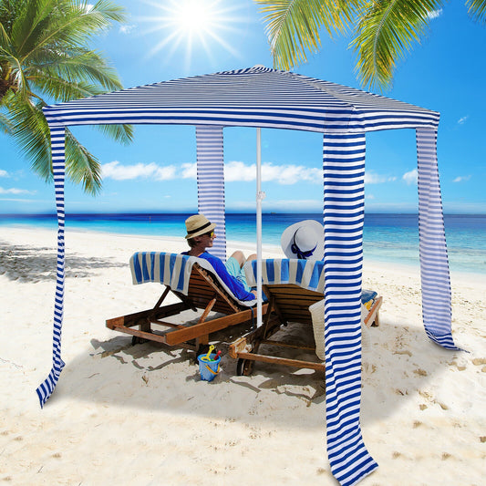 A navy and white striped beach canopy set up on a beach with a person lounging on a chair under it, surrounded by palm trees and the ocean in the background.