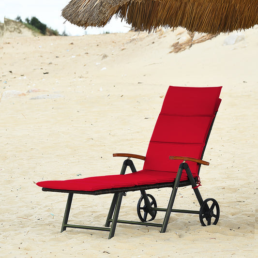 A red outdoor chaise lounge chair with wheels and a cushion, set on a sandy beach under a straw umbrella.