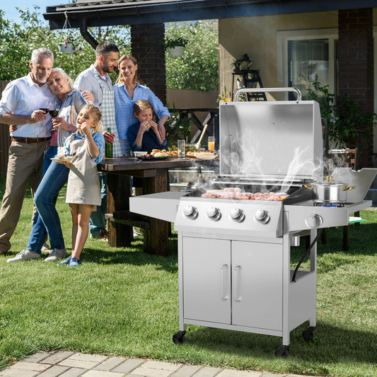 A silver propane gas grill with 5 burners and side burners, featuring a closed lid and side tables with grilling tools and food. A family is gathered around a table in the background, appearing to enjoy outdoor food prepared on the grill.