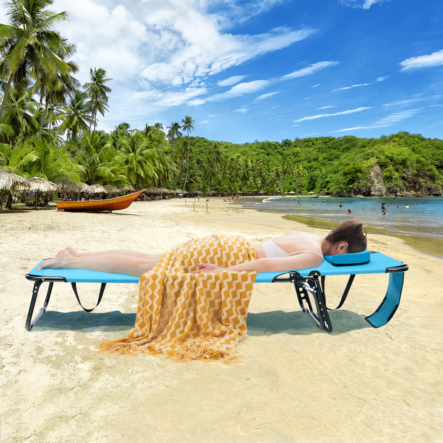 A person sunbathing on a blue beach chaise lounge chair with a face hole, located on a tropical beach with greenery and a boat in the background.