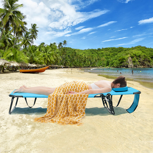 A person sunbathing on a blue beach chaise lounge chair with a face hole, located on a tropical beach with greenery and a boat in the background.