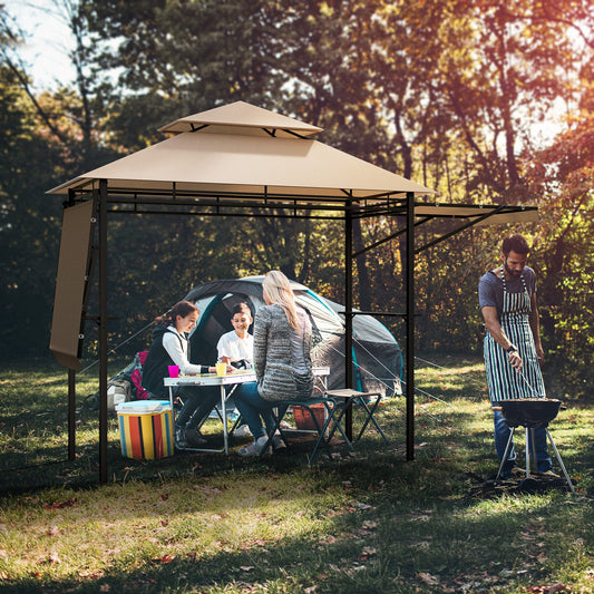 A beige patio BBQ grill gazebo with dual side awnings, set up outdoors with people in the background enjoying the space.