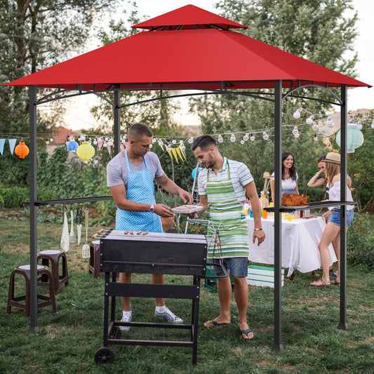 An outdoor barbecue grill gazebo with a red canopy, located in a garden setting with people gathered around the grill.
