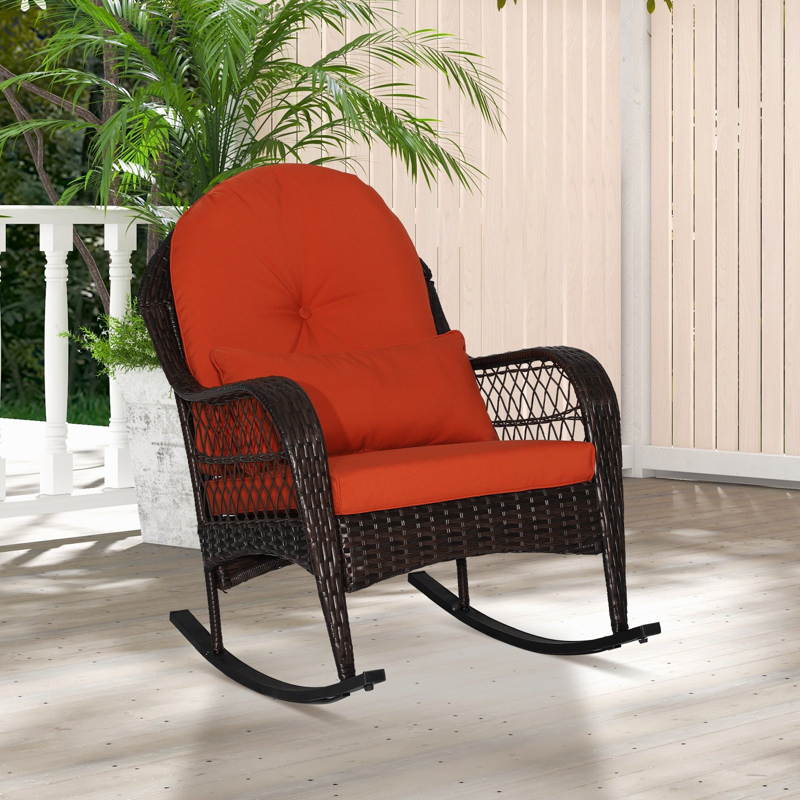 A red cushioned patio rocking chair made with a metal frame and PE rattan, displayed on a balcony with green plants in the background.