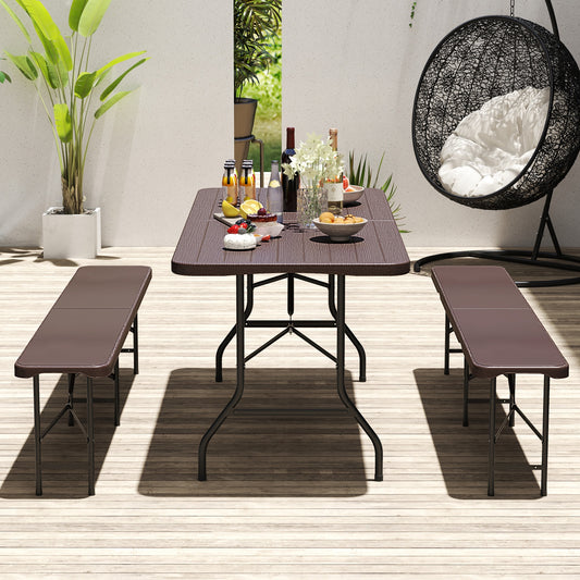 A brown folding table with wood grain HDPE tabletop and metal legs, set outdoors with a bench on each side and a variety of food on the table.