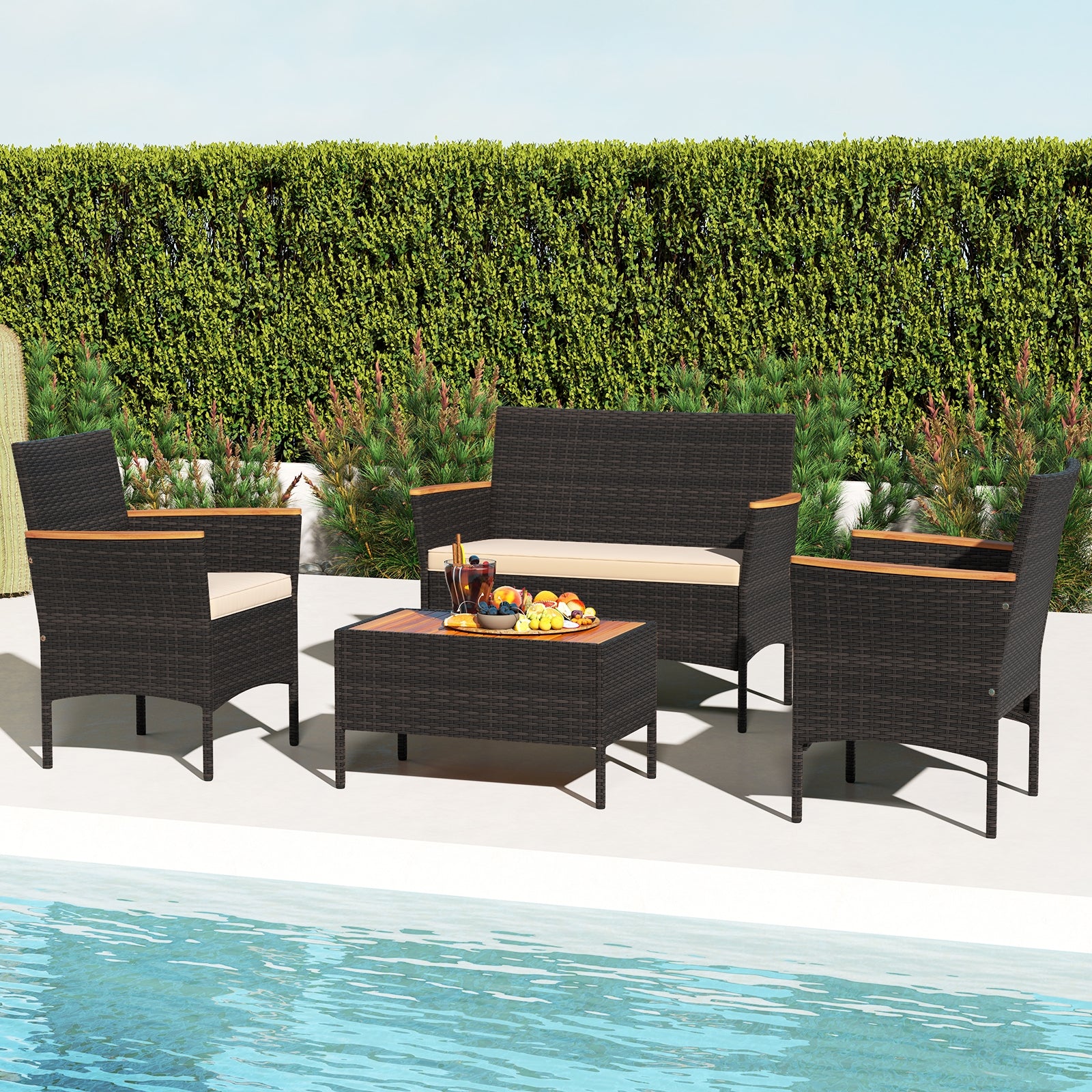A 4 piece patio wicker sofa set with two rattan chairs and a loveseat with a table placed by a poolside, with a hedge in the background.