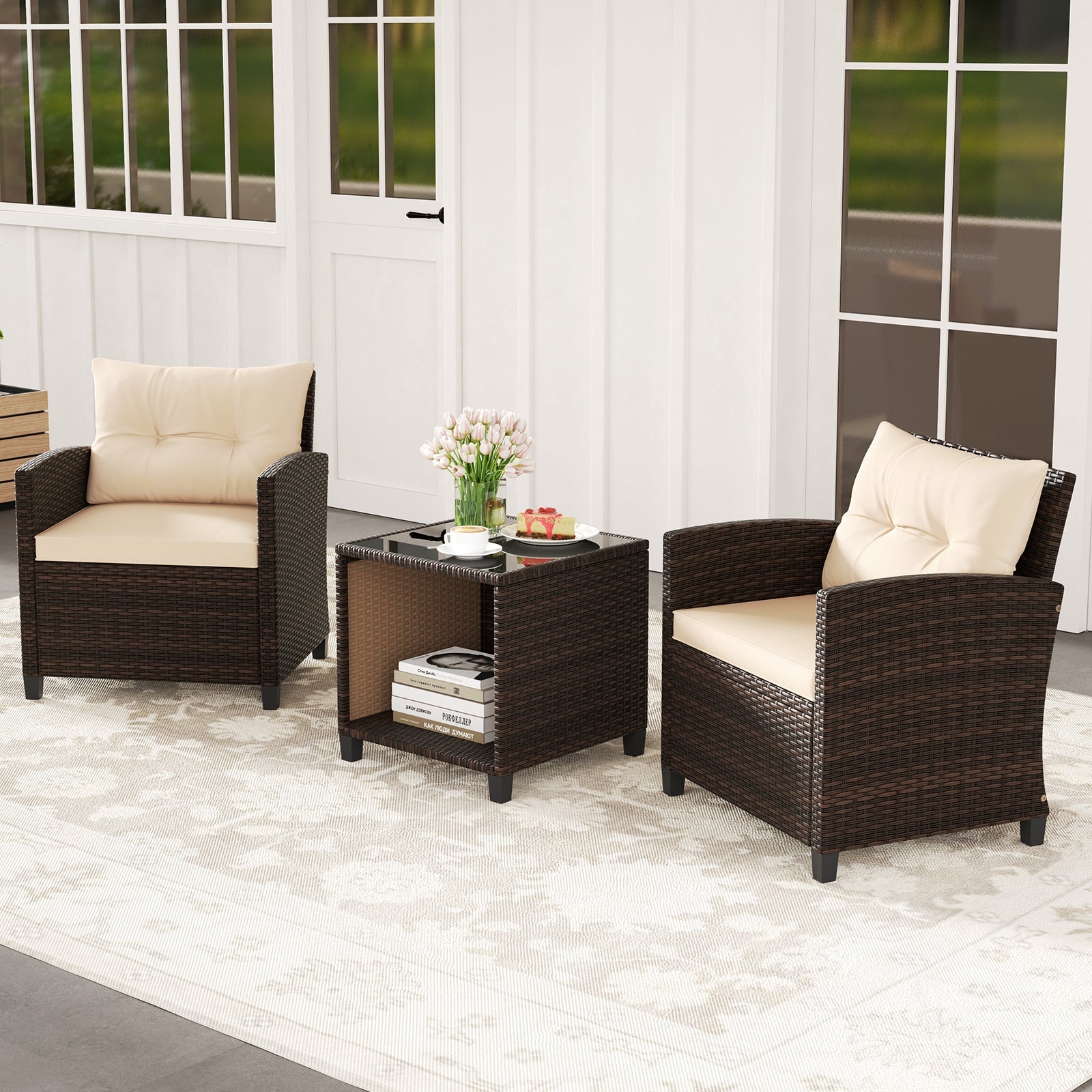 A three piece patio sofa set with two wicker chairs and a coffee table with a storage compartment. The set is beige and brown in color with cushions on the chairs and a book and a vase of flowers on the table, placed on a white rug.