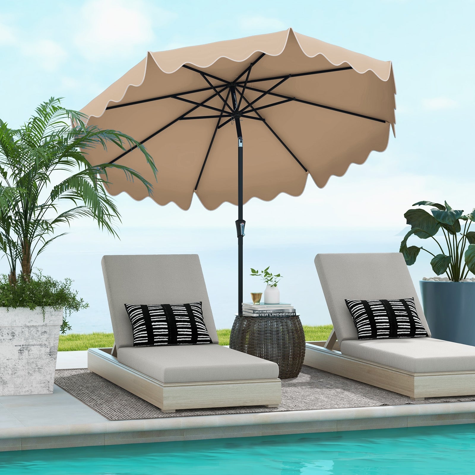A brown patio umbrella with a crank handle, placed over two beige lounge chairs by a pool, with decorative pillows, a side table, and potted plants in the background.