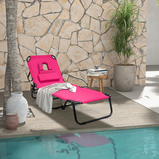 A pink beach chaise lounge chair with a face hole, displayed by a poolside with a stone wall background and some decorative plants.