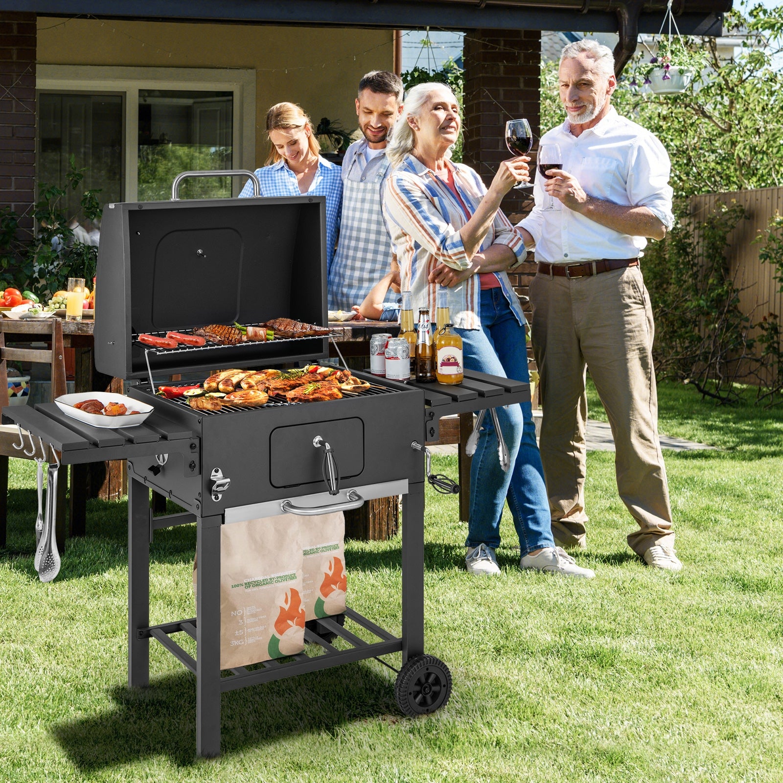 An outdoor BBQ charcoal grill with two foldable side tables and wheels, cooking food on the grill with people gathered around enjoying wine.