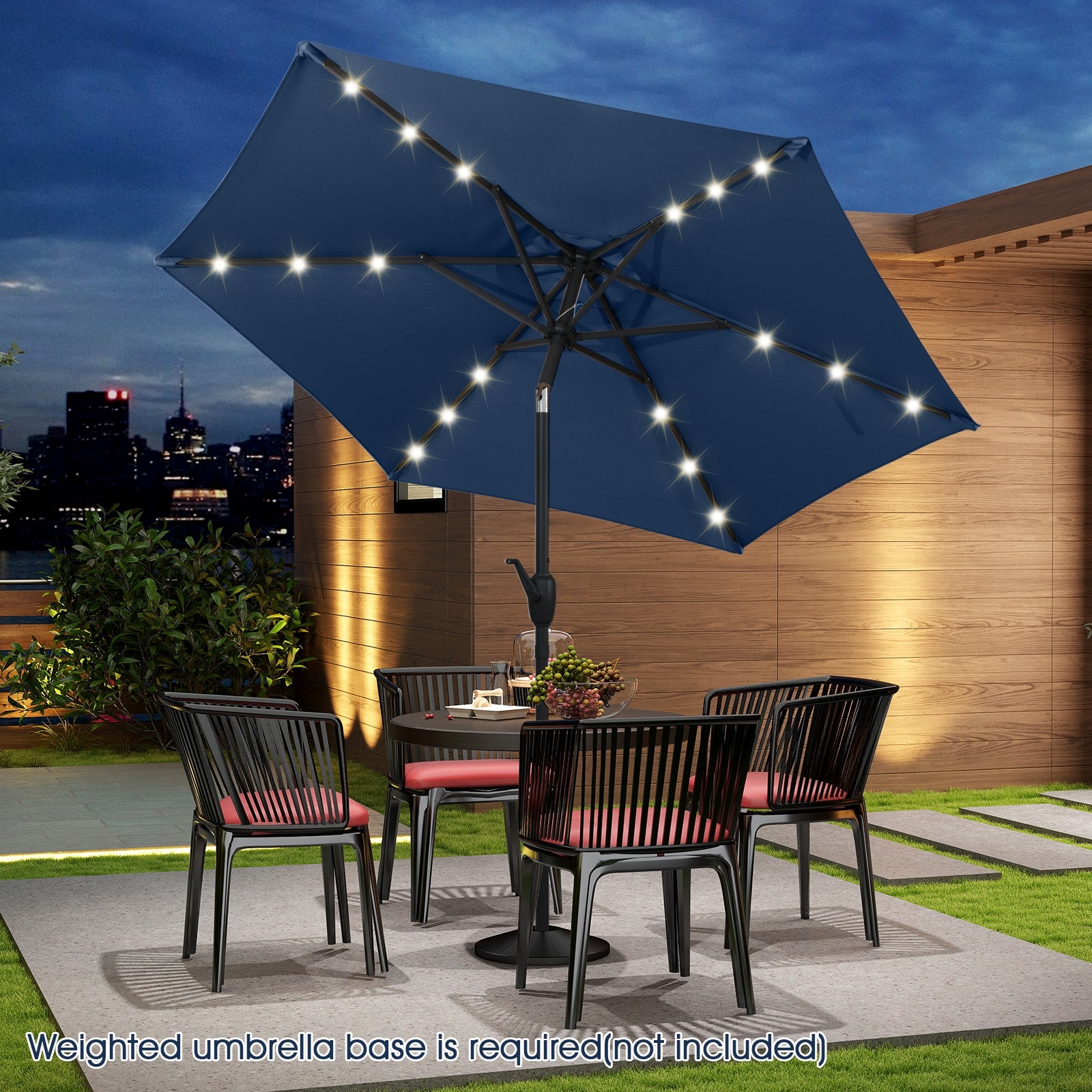 A navy blue patio umbrella with LED lights, featuring a crank handle, placed over a table and chairs set on a patio. The image is taken at dusk with a cityscape in the background.