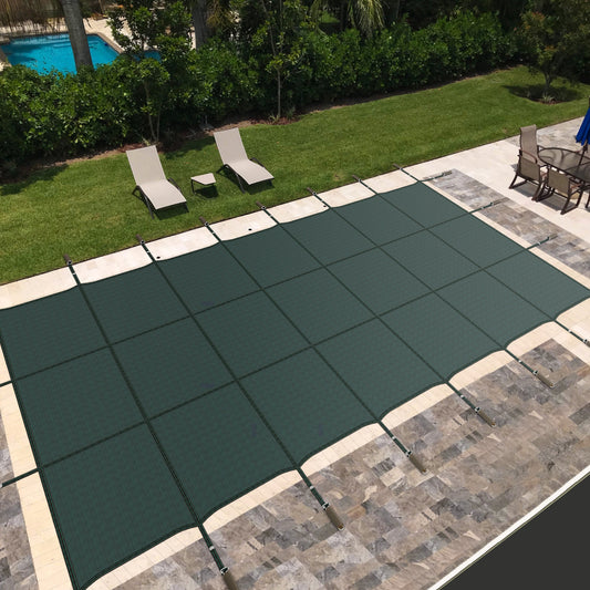 A green pool cover laid on an in-ground swimming pool, surrounded by a stone deck with lawn chairs and a pool umbrella in the background.