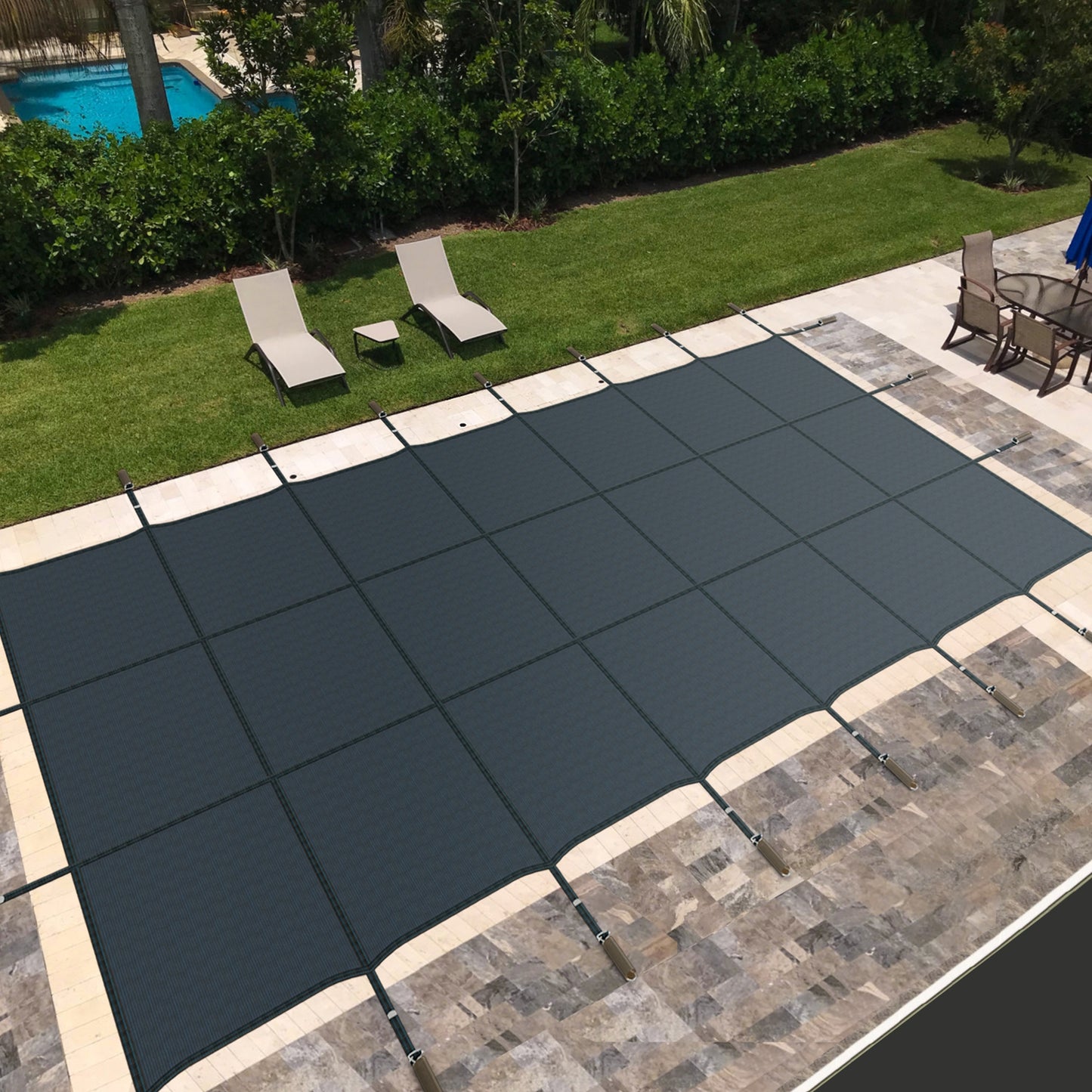 A blue safety pool cover installed on an in-ground swimming pool, surrounded by a stone deck with patio furniture in the background.