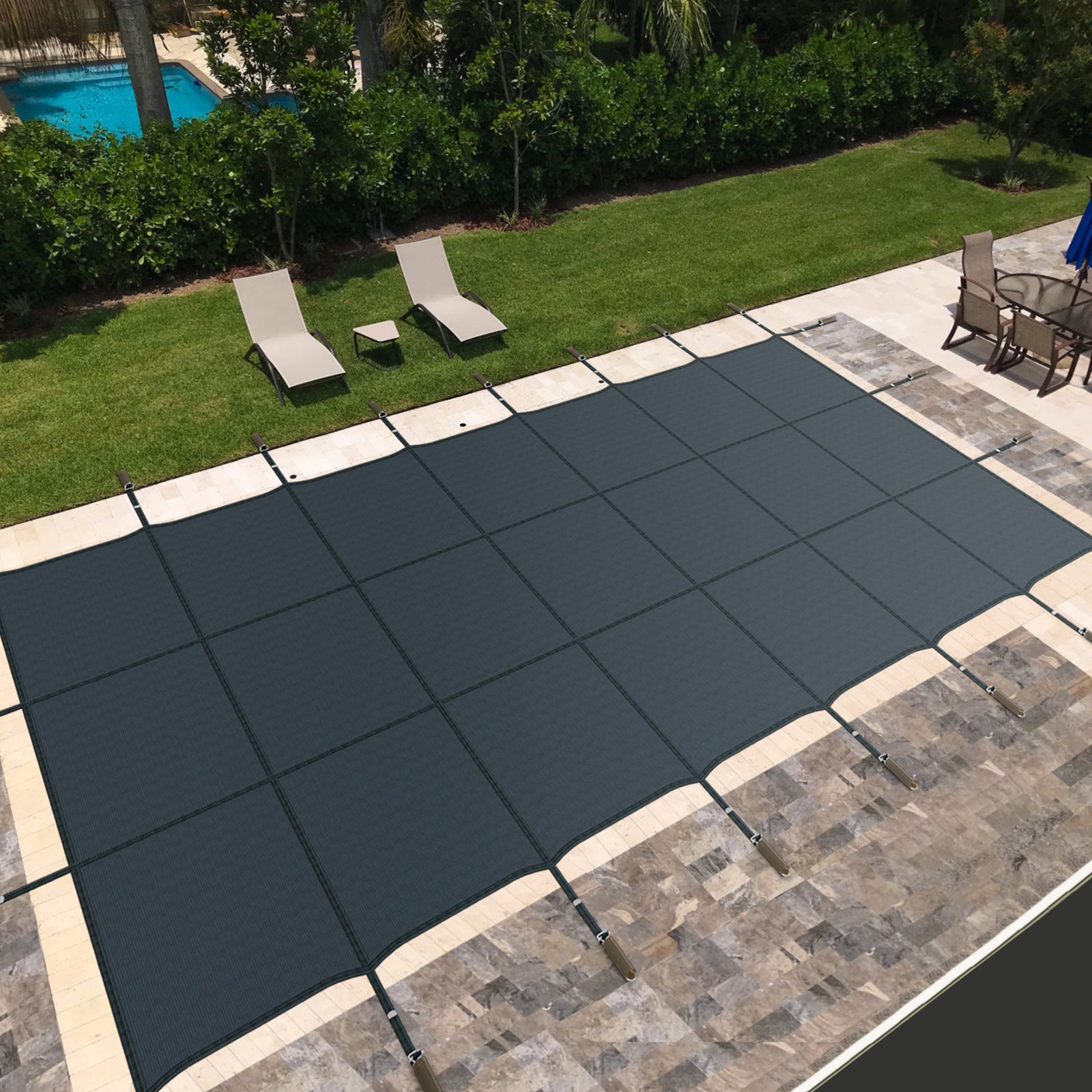 A blue safety pool cover installed on an in-ground swimming pool, surrounded by a stone deck with patio furniture in the background.